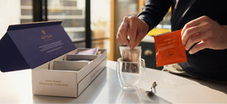 A woman hold an Artisan Coffee co coffee bag above a blue mug that is on top of some scales. To the left of this is a sugar bowl and a kettle